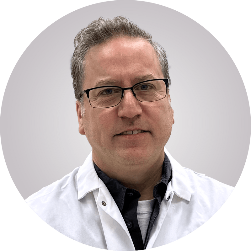 Close-up portrait of a middle-aged man with gray hair and black-framed glasses, wearing a white lab coat over a dark shirt, smiling subtly against a light gray circular background.